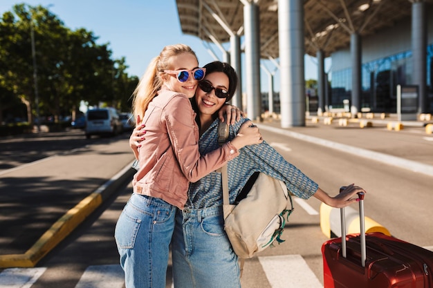 Two beautiful girls in sunglasses happily hugging each other while looking in camera with red suitcase and backpack on shoulder outdoor near airport