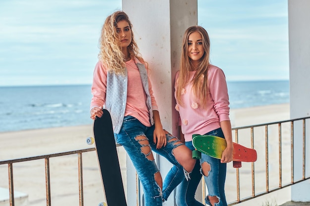 Foto gratuita due belle ragazze in abiti alla moda in posa con skateboard vicino a un guardrail contro una costa del mare.