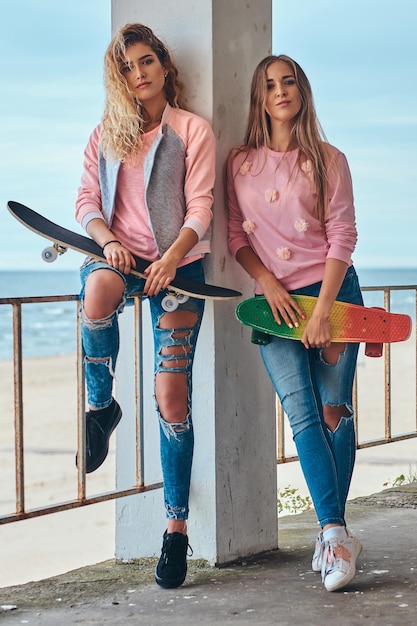Foto gratuita due belle ragazze in abiti alla moda in posa con skateboard vicino a un guardrail contro una costa del mare.