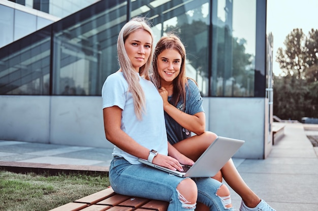 Two beautiful hipster girls sitting on the bench with a laptop on a background of the skyscraper.