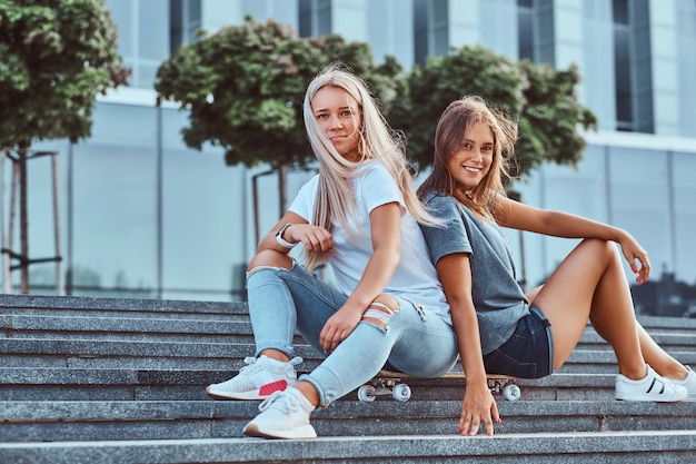 Две красивые хипстерские девушки сидят на ступеньках со скейтбордом на фоне небоскреба.