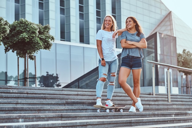 Two beautiful hipster girls standing on steps with skateboard on a background of the skyscraper.