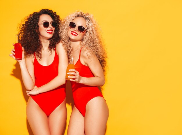 Две красивые сексуальные улыбающиеся женщины-хипстеры в красных летних купальных костюмах Модные модели с прической афро-кудри веселятся в студии Горячая женщина, изолированная на желтом в солнцезащитных очках