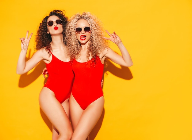 빨간 여름 수영복 수영복을 입은 아름다운 섹시한 웃는 힙스터 여성 두 명 아프로 컬 헤어스타일이 있는 최신 유행 모델 스튜디오에서 재미 있는 뜨거운 여성 노란색으로 격리