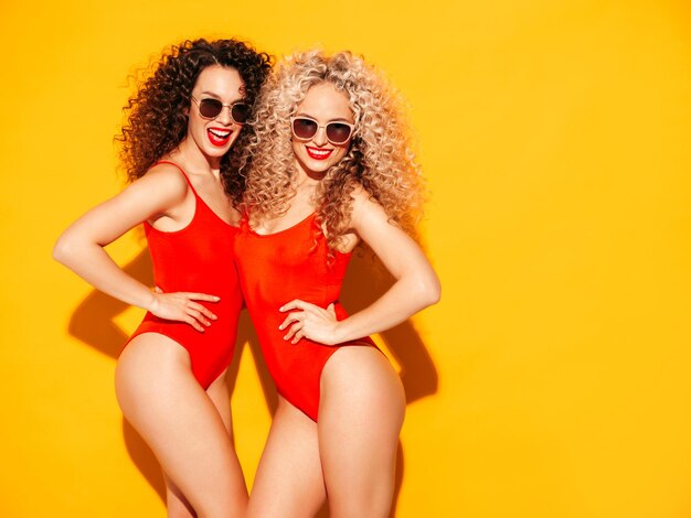 Two beautiful sexy smiling hipster women in red summer swimwear bathing suits Trendy models with afro curls hairstyle having fun in studio Hot female posing near yellow wall in sunglasses