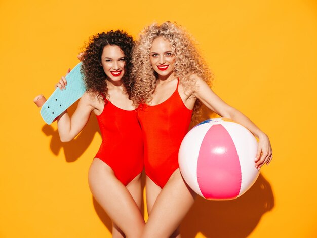 Две красивые сексуальные улыбающиеся женщины-хипстеры в красных летних купальных костюмах Модные модели с прической афро-кудри веселятся в студииГорячая женщина, изолированная на желтомХолдинг пенни скейтборд