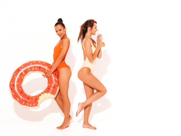 Two beautiful sexy smiling women in summer colorful swimwear bathing suits. Girls isolated. Funny models drinking fresh cocktail smoozy drink with donut lilo inflatable mattress