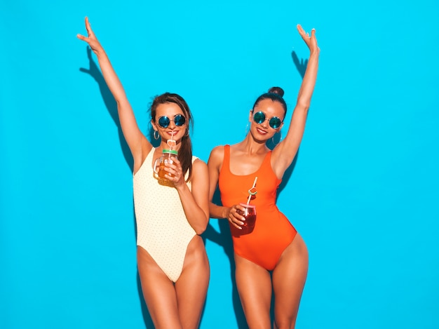 Две красивые сексуальные улыбающиеся женщины в летних разноцветных купальниках. Модные девушки в солнечных очках. Схожу с ума. Смешные модели изолированы. Пить свежий коктейль смузи напиток. Поднять руки