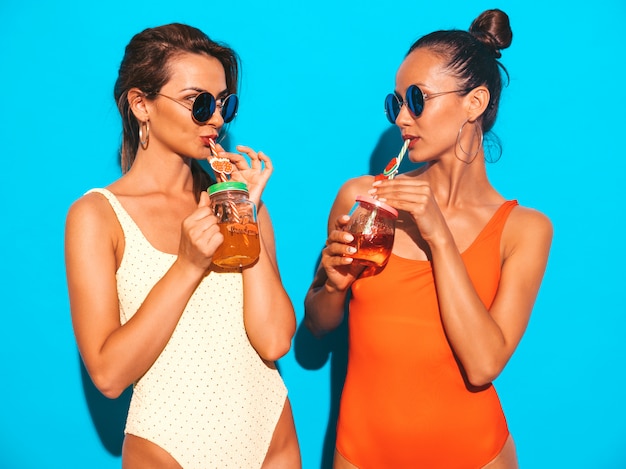 Две красивые сексуальные улыбающиеся женщины в летних разноцветных купальниках. Модные девушки в солнечных очках. Схожу с ума. Смешные модели изолированы. Пить свежий коктейль смузи напиток