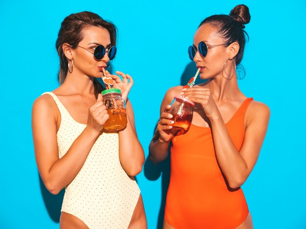 Two beautiful sexy smiling women in summer colorful swimwear bathing suits. Trendy girls in sunglasses. Going crazy. Funny models isolated. Drinking fresh cocktail smoozy drink