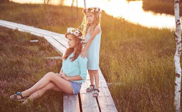 Two beautiful sisters standing against the background of a beautiful landscape, walk on the field near a pond at bright sunset.