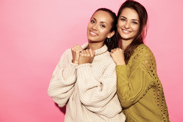 Due belle donne bellissime sorridenti. donne in piedi in eleganti maglioni bianchi e verdi, sul muro rosa.