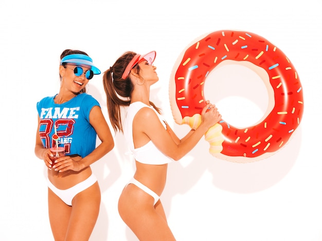 Free photo two beautiful smiling sexy women in white summer underpants and topic. girls in sunglasses,transparent visor cap. models drinking fresh cocktail smoozy drink with donut lilo inflatable mattress