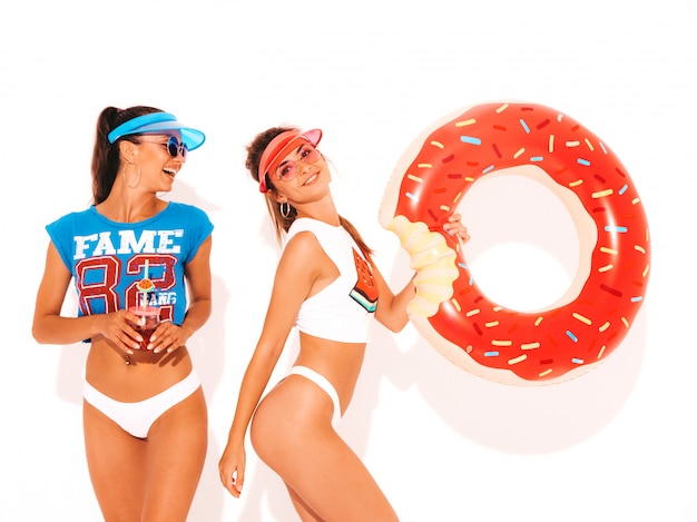 Free photo two beautiful smiling sexy women in white summer underpants and topic. girls in sunglasses,transparent visor cap. models drinking fresh cocktail smoozy drink with donut lilo inflatable mattress