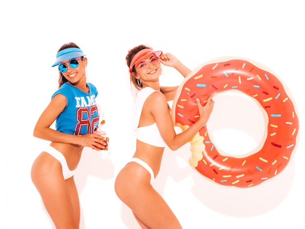 Free photo two beautiful smiling sexy women in white summer underpants and topic. girls in sunglasses,transparent visor cap. models drinking fresh cocktail smoozy drink with donut lilo inflatable mattress