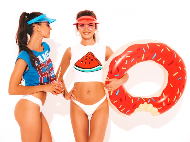 Free photo two beautiful smiling sexy women in white summer underpants and topic. girls in sunglasses,transparent visor cap. models drinking fresh cocktail smoozy drink with donut lilo inflatable mattress