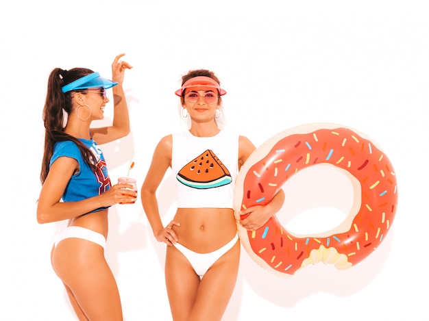 Two beautiful smiling sexy women in white summer underpants and topic. Girls in sunglasses,transparent visor cap. Models drinking fresh cocktail smoozy drink with donut lilo inflatable mattress