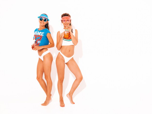 Two beautiful smiling sexy women in white summer underpants and topic. Trendy girls in sunglasses,transparent visor cap. Going crazy. Funny models isolated. Drinking fresh cocktail smoozy drink