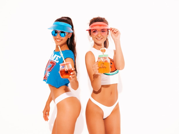 Free photo two beautiful smiling sexy women in white summer underpants and topic. trendy girls in sunglasses,transparent visor cap. going crazy. funny models isolated. drinking fresh cocktail smoozy drink