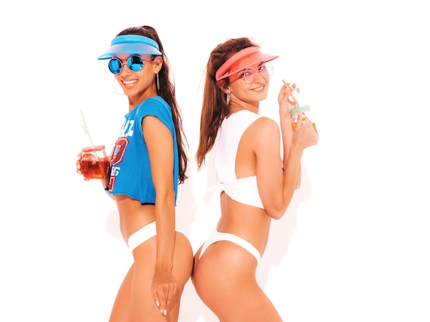 Free photo two beautiful smiling sexy women in white summer underpants and topic. trendy girls in sunglasses,transparent visor cap. going crazy. funny models isolated. drinking fresh cocktail smoozy drink