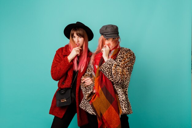 Две красивые женщины в стильных шубах из искусственного меха и шерстяном шарфе позируют на бирюзовой стене