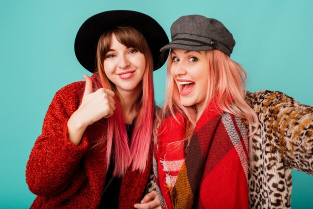 Two beautiful women in stylish faux fur coats and wool scarf posing on turquoise wall