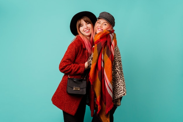 Two beautiful women in stylish faux fur coats and wool scarf posing on turquoise wall