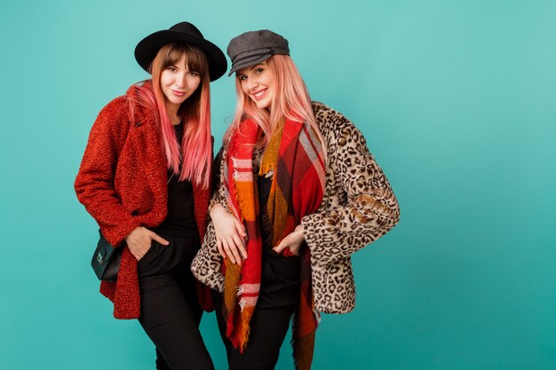 Two beautiful women in stylish faux fur coats and wool scarf posing on turquoise wall