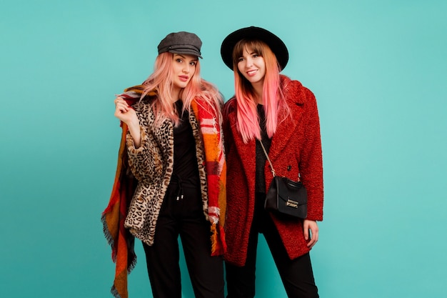 Two beautiful women in stylish faux fur coats and wool scarf posing on turquoise wall