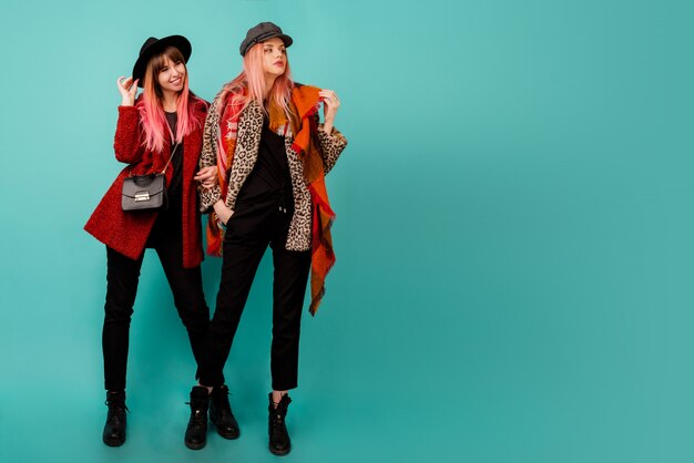 Two beautiful women in stylish faux fur coats and wool scarf posing on turquoise wall