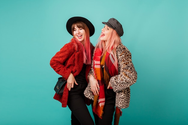 Free photo two beautiful women in stylish faux fur coats and wool scarf posing on turquoise wall