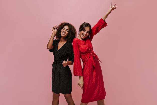 Two beautiful women with brunette hair in fashionable black and red dresses smiling and having fun on pink isolated backdrop