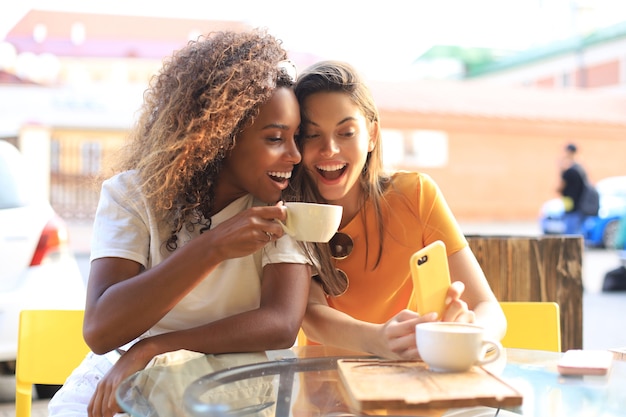 コーヒーを飲みながら携帯電話を見ているカフェに座っている2人の美しい若い女性。