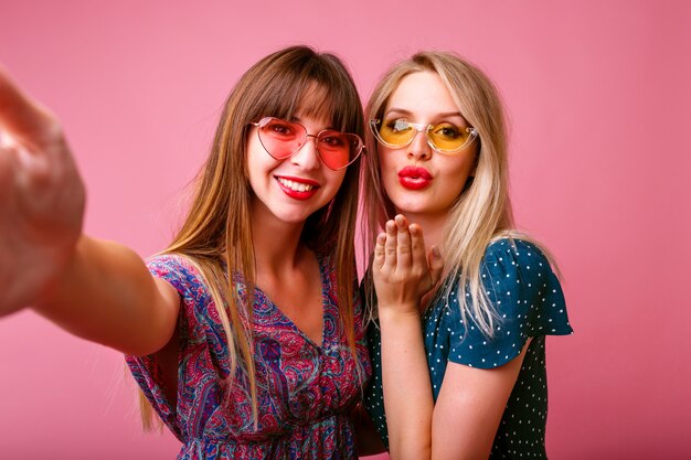 Two best friends sister women making selfie at pink wall, sending air kisses and smiling, stylish dresses and sunglasses, spring summer mood.