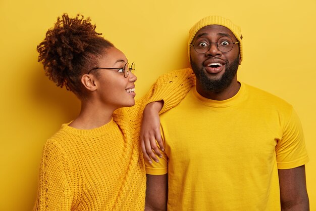 Two black friends in yellow clothes, have joyful looks, African American woman leans on shoulder of bearded guy