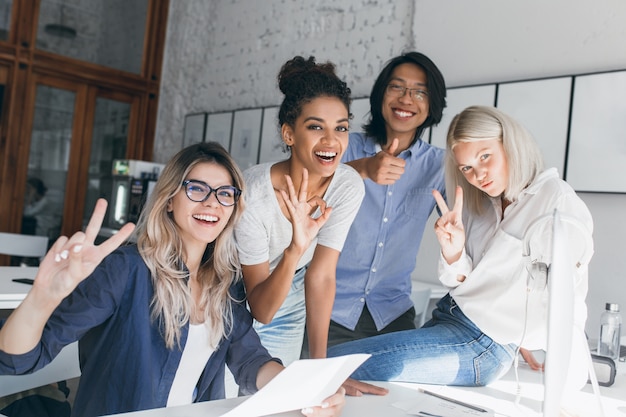 Two blonde female office workers posing with peace sign while fooling around with foreign colleagues