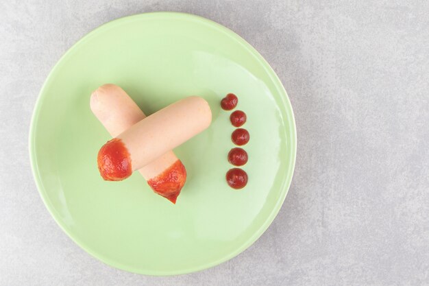 Two boiled sausages with ketchup on green plate.