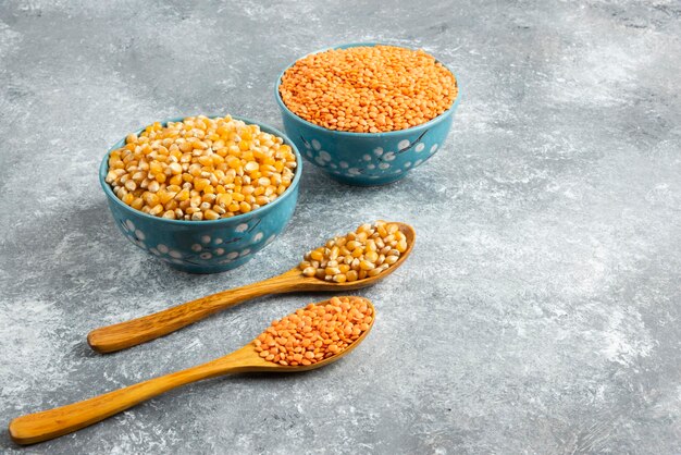 Two bowls of corn kernels and red lentil on marble surface.