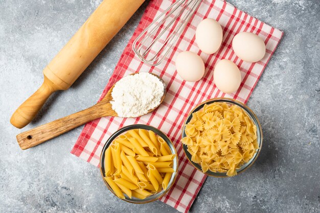 Two bowls of raw pasta, eggs, spoon of flour and rolling pin on marble table with tablecloth. 