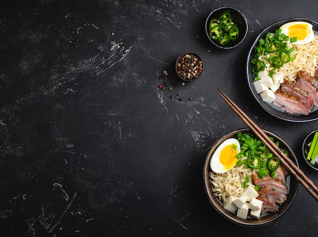 Two bowls of tasty asian noodle soup ramen with broth  tofu  pork  egg on black rustic stone background  space for text  close up  top view. hot tasty japanese ramen soup for dinner with copy space