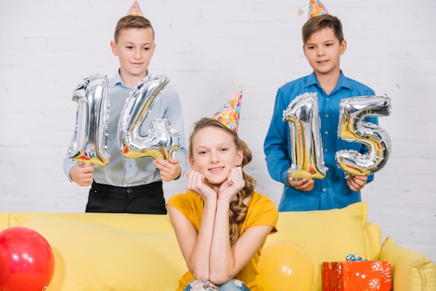 Foto gratuita due ragazzi in possesso di numeri 14 e 15 palloncini foil in mano in piedi dietro la ragazza di compleanno