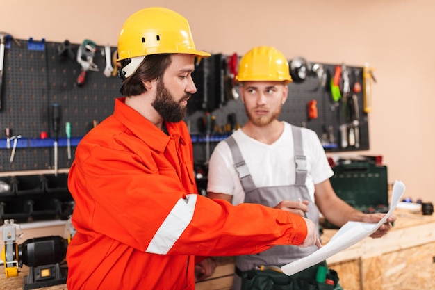 Two builders in work clothes and yellow hardhats working in workshop with tools on background