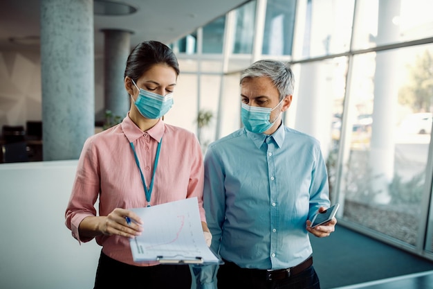 Two business people wearing face masks while examining paperwork in a hallway