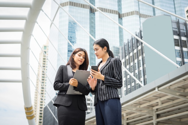 Two business woman standing using smartphone and discussing in front of the office. Business working concept.