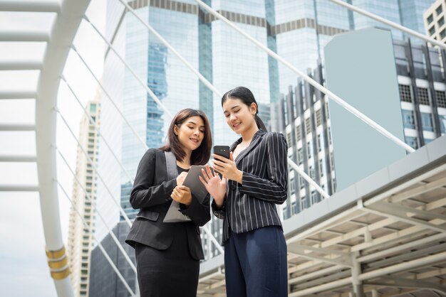 Two business woman standing using smartphone and discussing in front of the office. Business working concept.
