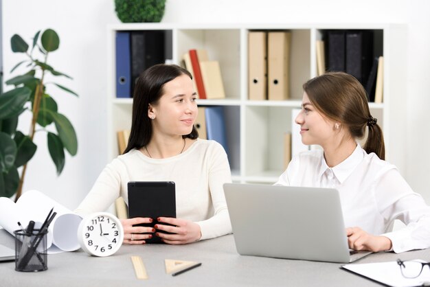 Two businesswoman holding digital tablet and laptop looking at each other