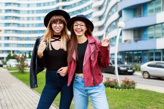 Two carefree smiling women posing  on modern city. Wearing wool hat leather jacket and jeans.