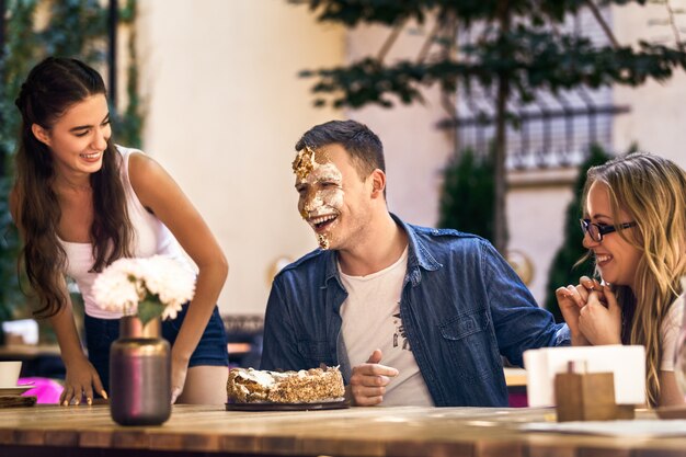 Two caucasian girls and a guy with face diry with cake cream are laughting and sitting around the table outdoors