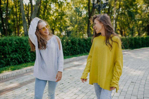 Two charming European women with long hairstyle in bright outfits are walking in the park and smiling