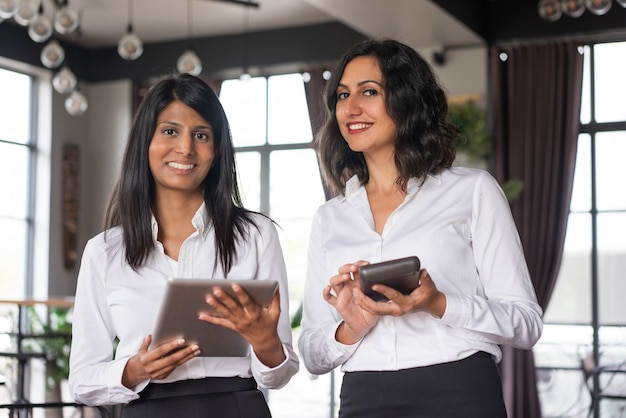 Two cheerful female colleagues using gadgets in cafe.